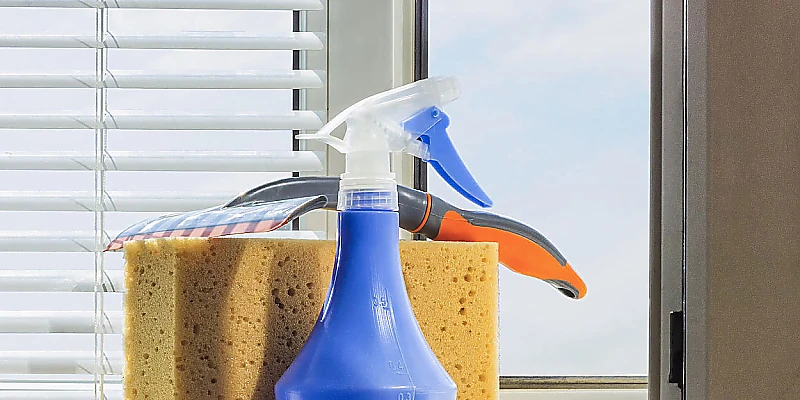 Set aus Fensterreinigungszubehör mit Sprühflasche, Schwamm und Abzieher vor Jalousie und Fenster.